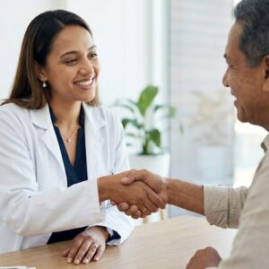 A doctor and patient shake hands warmly in a bright, modern medical consultation room.